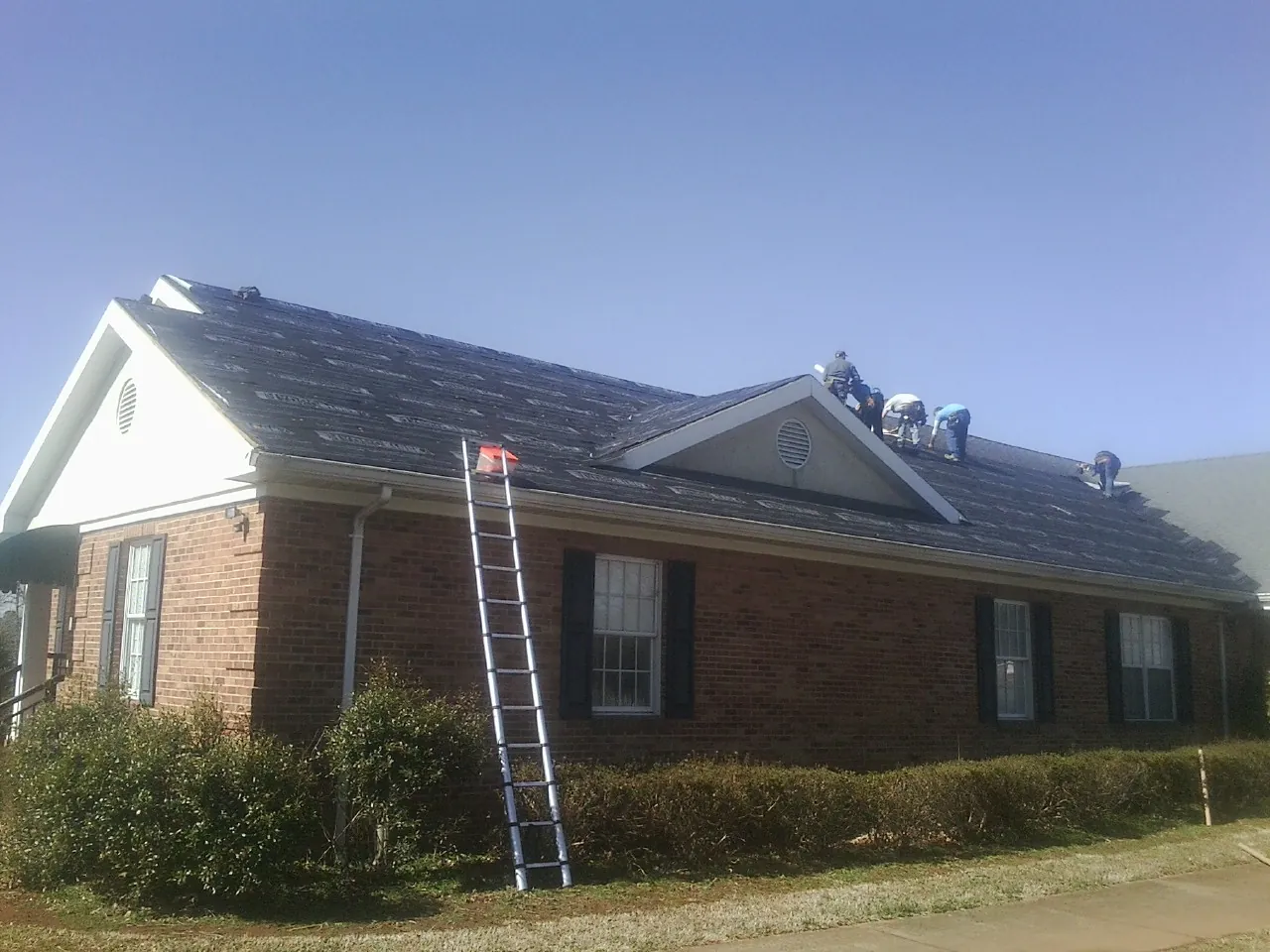 Crew tearing shingles off a brick ranch roof with a ladder set — Emergency Roof Repair work by our crew in Lowden