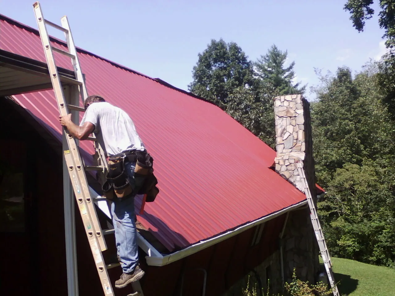 Roofer on a ladder beside a red metal roof and stone chimney — Soffit & Fascia Repair by Roofing Brothers in Lowden